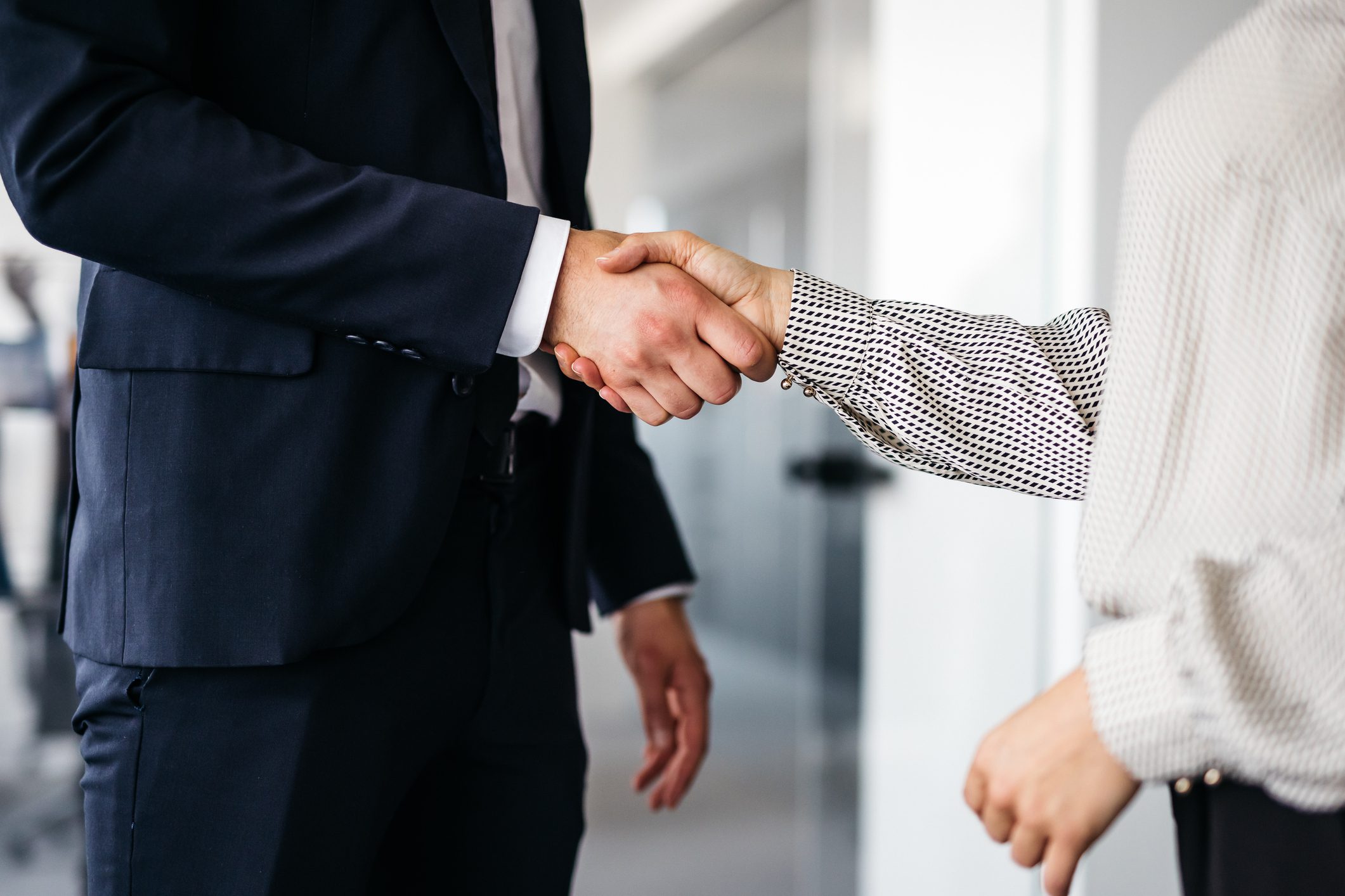 A close-up shot of two smartly dressed business co-workers shaking hands at work. Their faces are not visible. Horizontal daylight indoor photo.